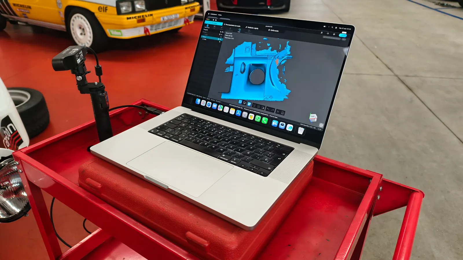 MacBook con el software de escaneado 3D mostrando la malla de una pieza del Porsche 924, con el escáner de mano y un clásico de rallye al fondo